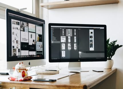 Dual monitor setup displaying good web design layouts and wireframes on a modern desk, showcasing UI/UX planning tools, with a notepad, phone, and decorative items nearby